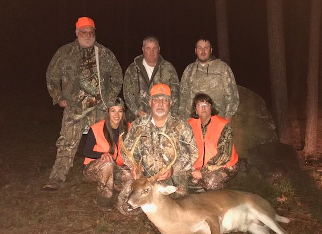 Six hunters in camouflage pose at night with a deer on the ground and a flashlight-lit forest behind them