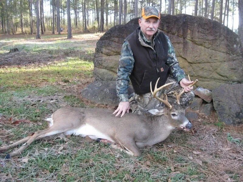 Hunter kneels behind a large buck deer in a wooded clearing, posing with the animal.