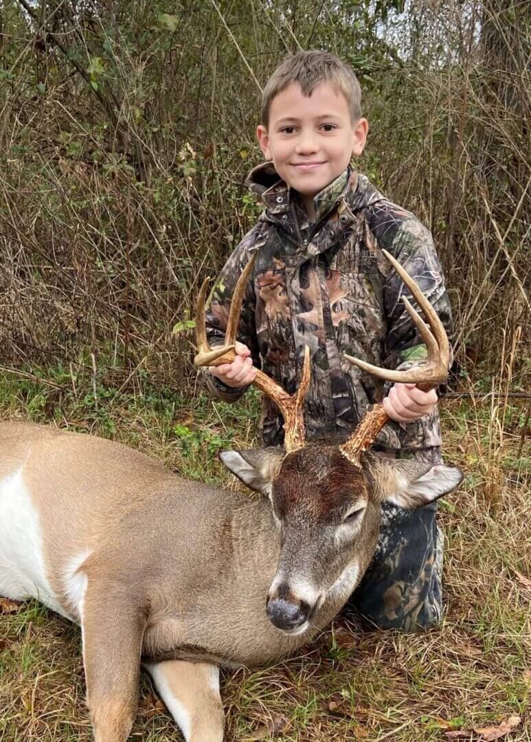 Boy standing beside a deer he is holding by the antlers in a wooded area