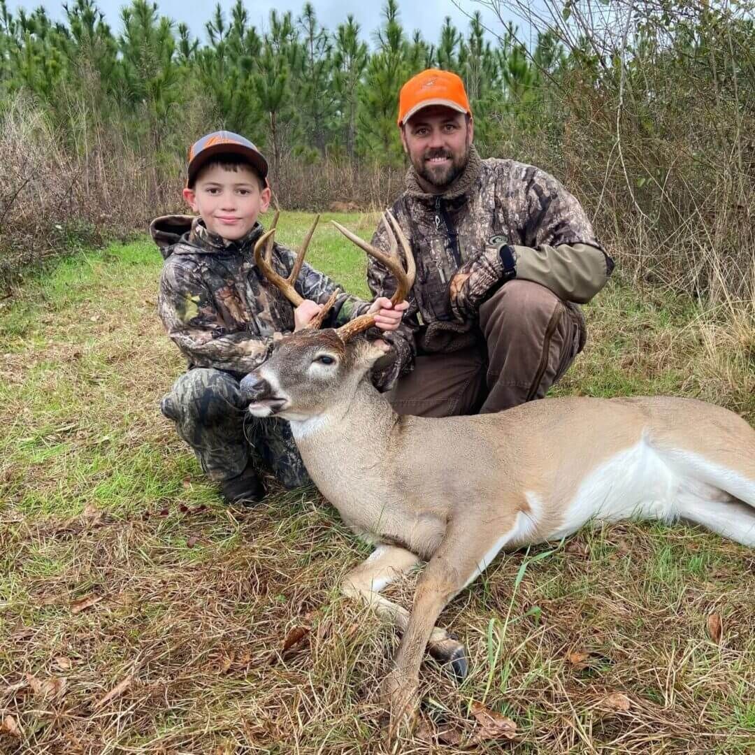 Adult and child posing with a deer in a grassy field, both wearing camouflage and orange caps.