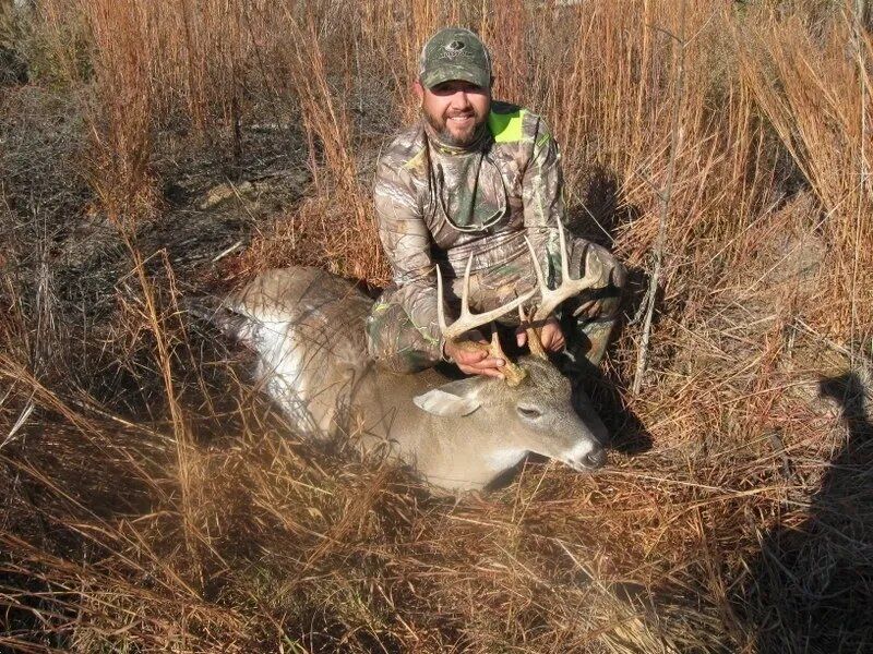Hunter kneels beside a deer in tall dry grass, holding its antlers in a marshland.