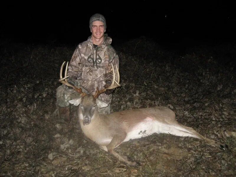 Hunter kneels beside a deer at night, holding the animal’s antlers in a dark field.