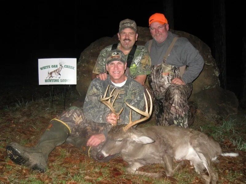 Three hunters pose with a deer and antlers at night beside a hunting sign.