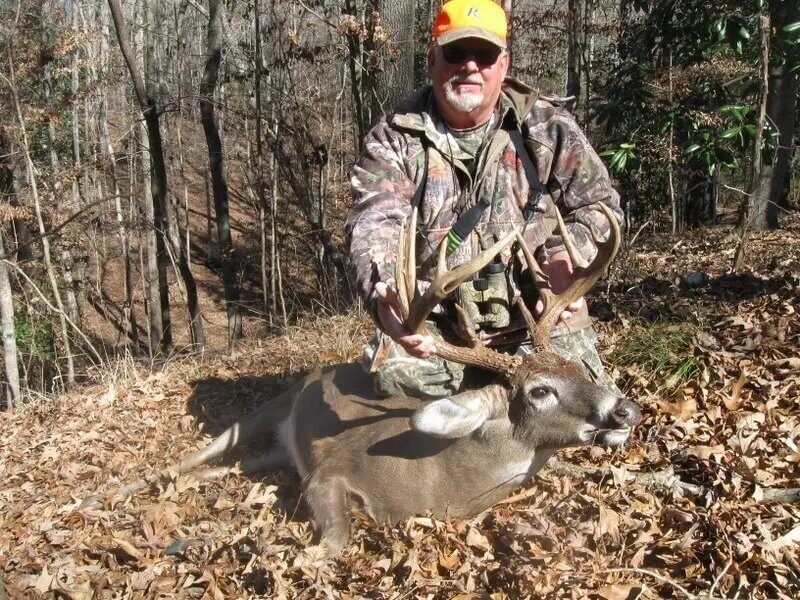 Hunter in camouflage holding a deer in a wooded area, wearing an orange cap and sunglasses.