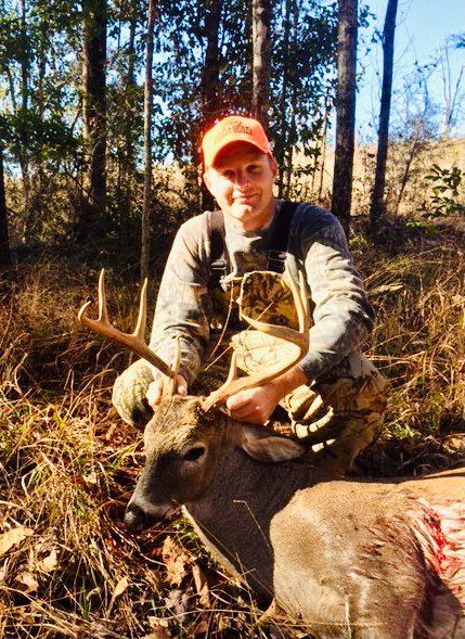 Hunter in orange cap kneels beside a deer in a wooded area, holding the antlers.