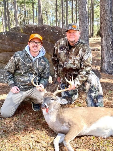 Two hunters kneeling beside a deer in a wooded area, wearing camouflage and orange caps