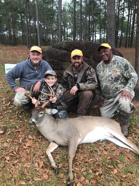 Four hunters posing with a deer in a pine forest, one holding a young child.