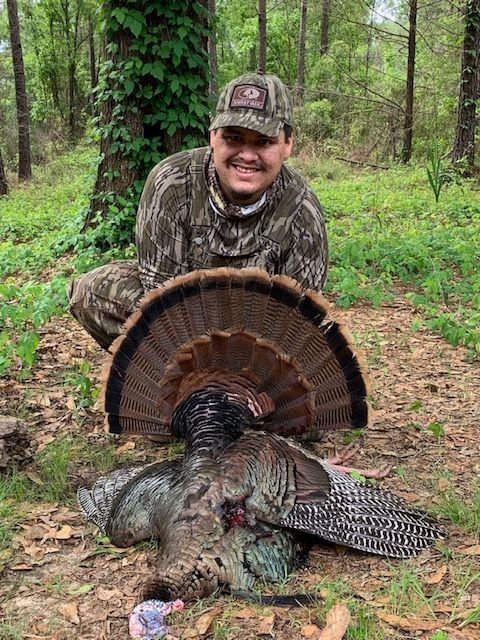 Hunter kneeling behind a wild turkey in a wooded area, smiling at the camera.