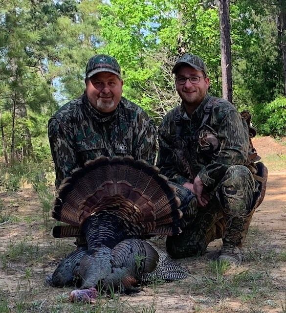 Two hunters pose with a wild turkey in a wooded clearing