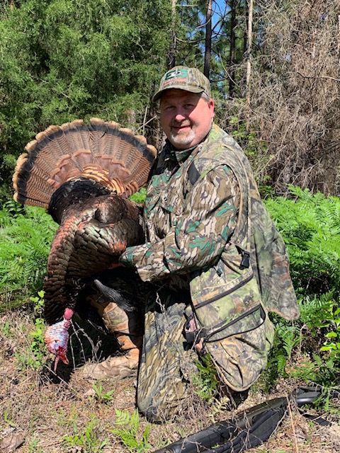 Hunter kneels beside a turkey in a forest clearing, wearing camouflage with the bird’s tail fanned.