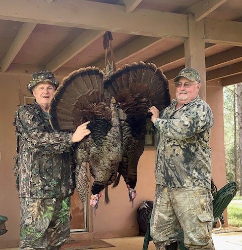 Two camouflaged hunters holding a large turkey by its tail under a porch roof