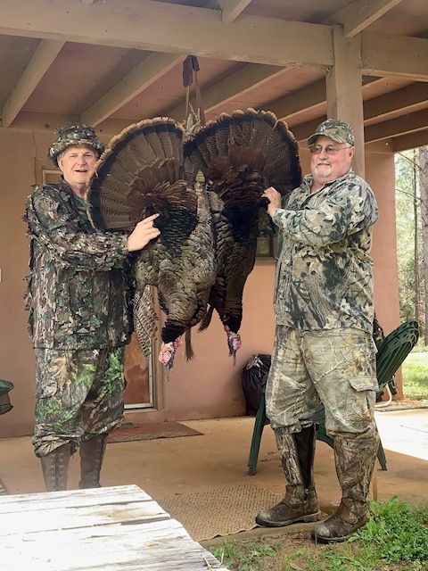 Two hunters holding a large turkey by the legs on a porch under a wooden roof.