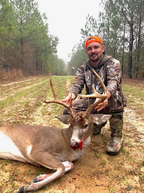 Hunter kneels beside a buck in a forest clearing, holding its antlers and smiling.