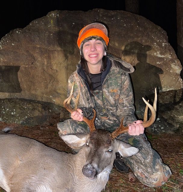Hunter in camouflage kneels with a deer at night, wearing an orange hat beside a large rock.