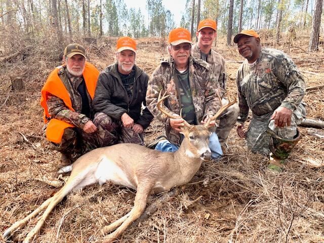 Five hunters posing with a deer in a wooded field, wearing orange and camouflage