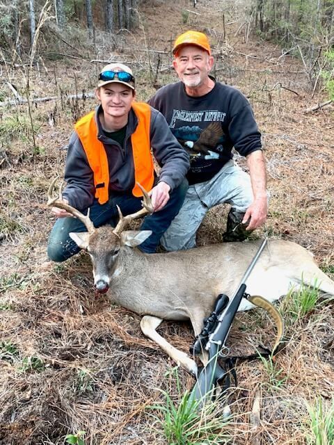 Two hunters kneel beside a deer in a grassy field, one in orange vest and both holding the antlers.