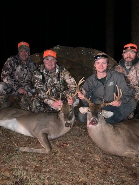 Four hunters pose with two deer at night, wearing camouflage and orange caps.