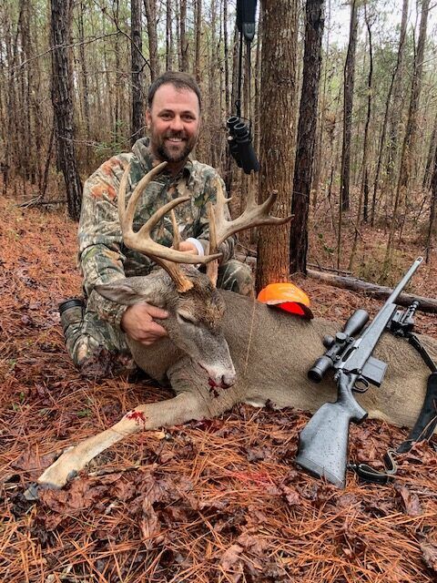 Hunter kneeling beside a large deer in a wooded area, with a rifle and orange cap nearby.