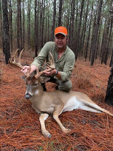 Hunter kneels beside a deer in a pine forest, holding its antlers; orange cap and camo shirt.