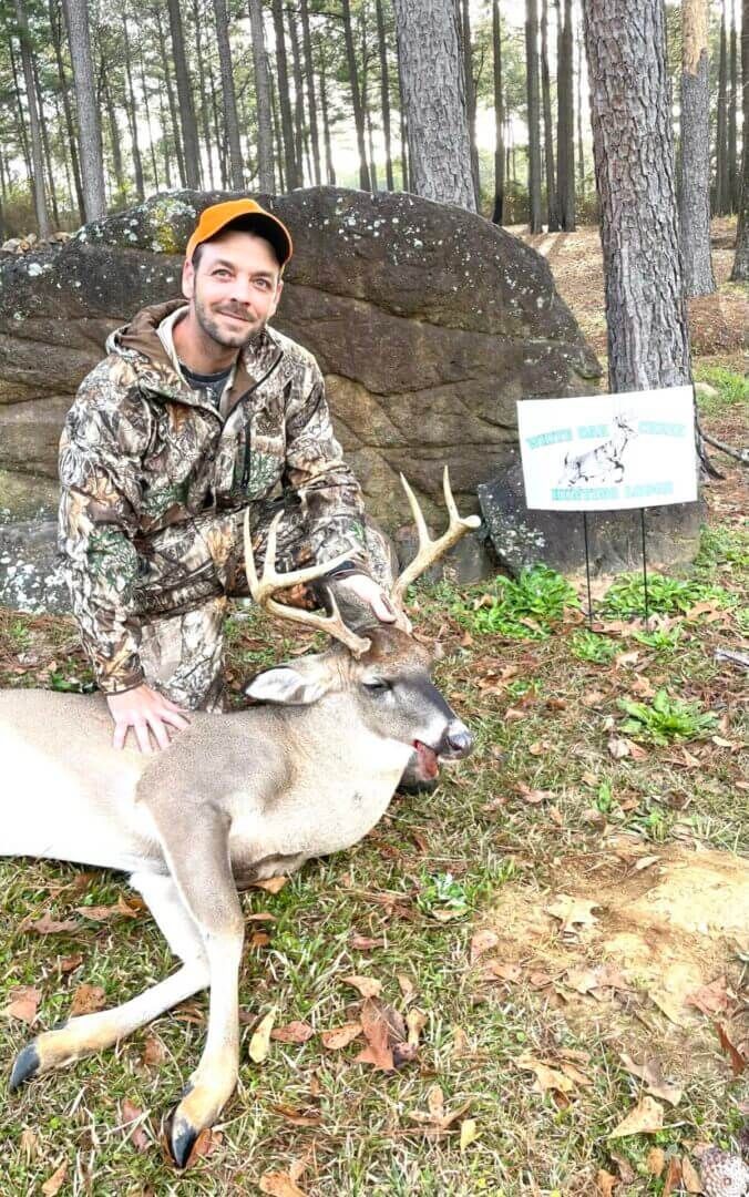 Hunter kneels beside a deer in a wooded area, with a sign posted on a rock behind him.
