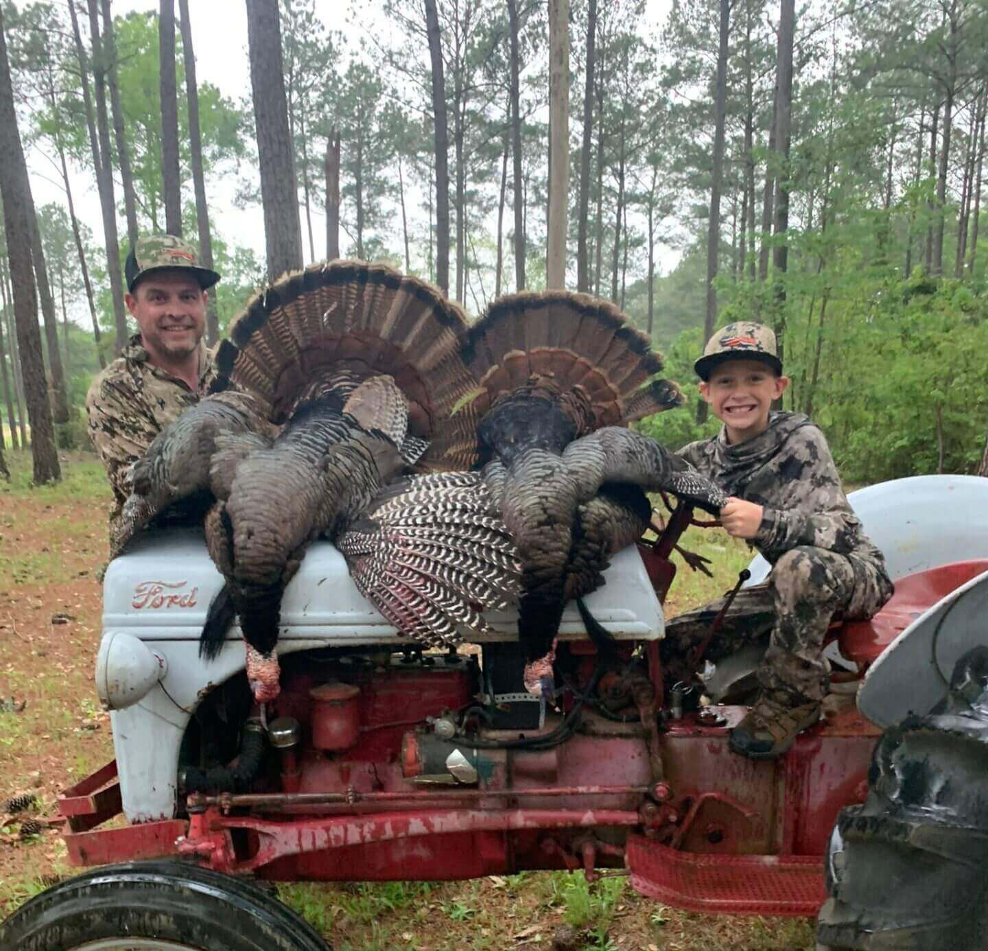 Two people posing with two turkeys on a red tractor in a wooded area
