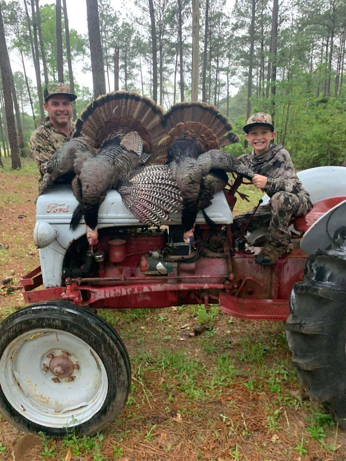 Two hunters pose with wild turkeys on a red tractor in a wooded field.