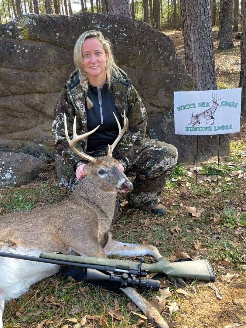 Hunter kneeling behind a deer with a rifle, posing beside a rock in a wooded area.