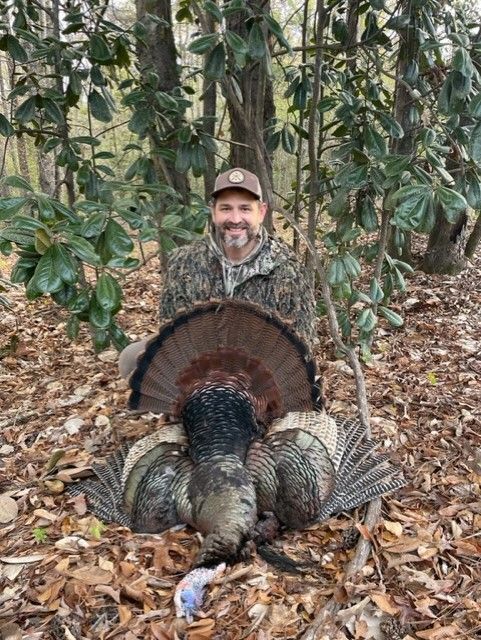 Hunter kneels beside a large wild turkey in a wooded area with fallen leaves.