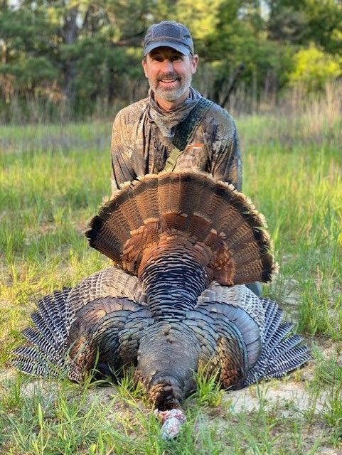 Hunter kneels behind a large wild turkey in a grassy field at sunset.