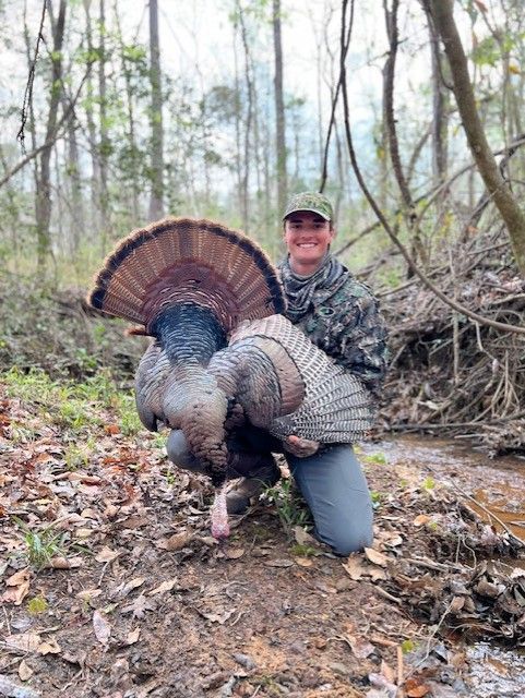Hunter kneeling with a large turkey in a wooded area, holding its fan spread wide