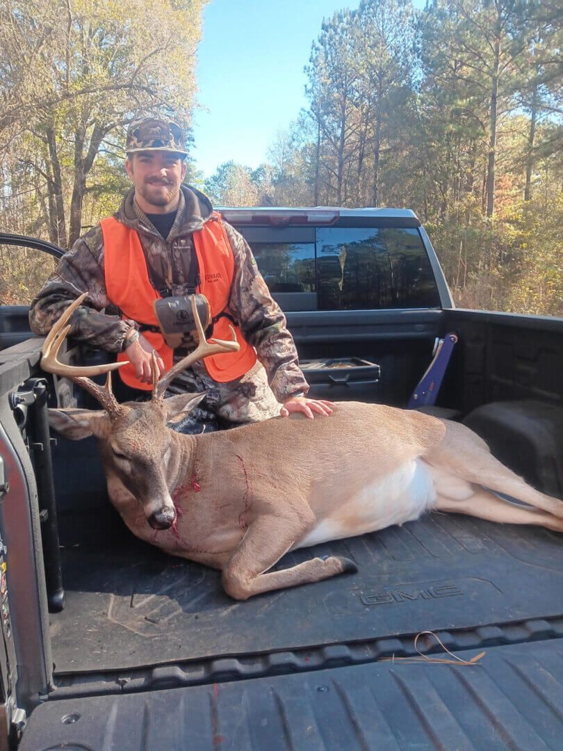 Hunter in orange vest posing with a deer in the back of a pickup truck in a wooded area.