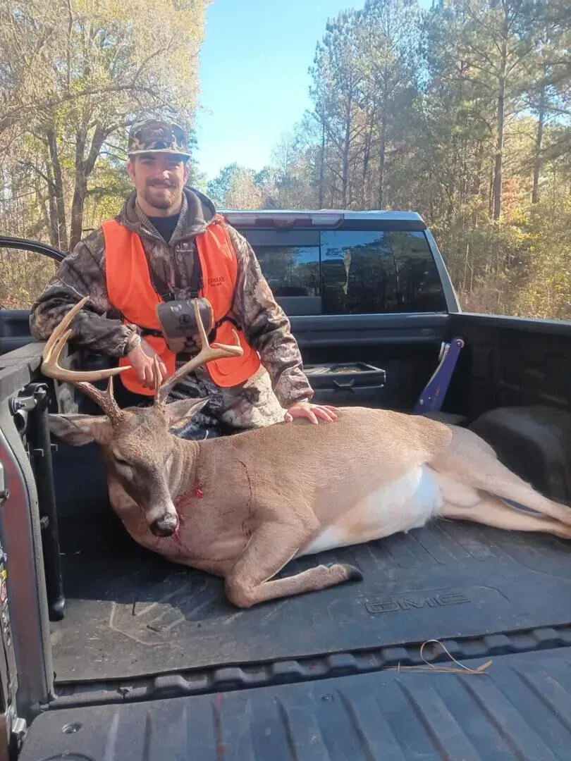 Hunter kneels in a pickup truck bed beside a buck, wearing an orange vest in a wooded area.