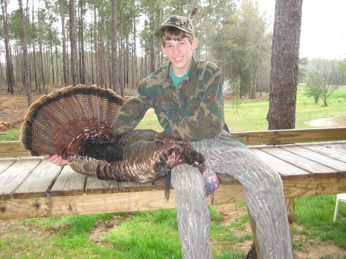 Hunter posing with a large turkey over a wooden bench in a grassy, wooded area