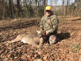 Hunter in camouflage kneels beside a deer in a wooded area, wearing an orange cap.