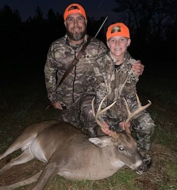 Two hunters in camouflage walking through a grassy field with rifles over their shoulders