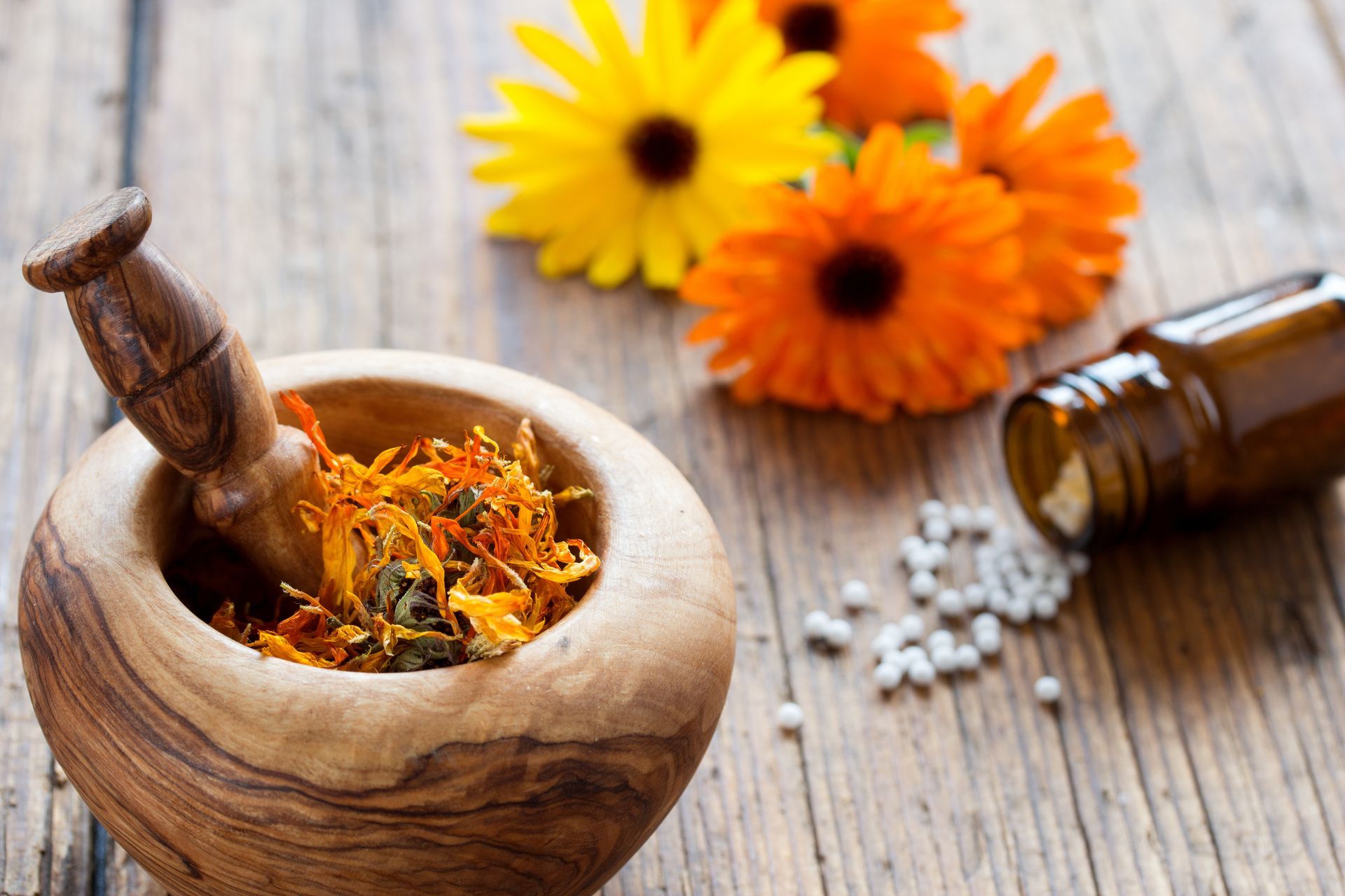 Mörser und Stößel mit Kräutern, Blumen und verschütteten Kapseln auf einem rustikalen Holztisch