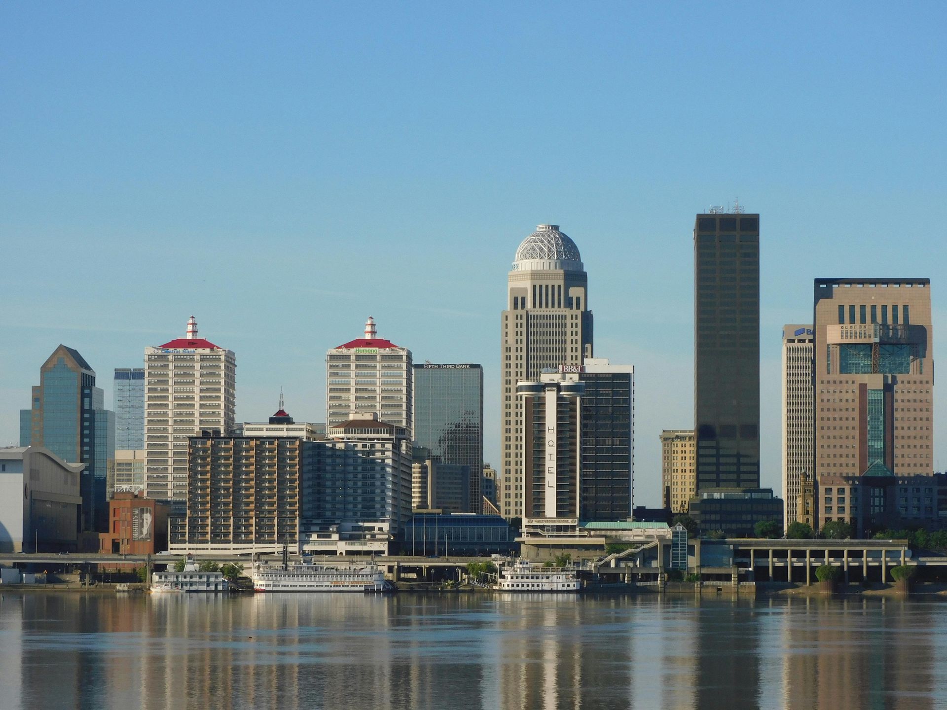 Skyline of Louisville, Kentucky, with buildings of varying heights reflected in the Ohio River under a blue sky.