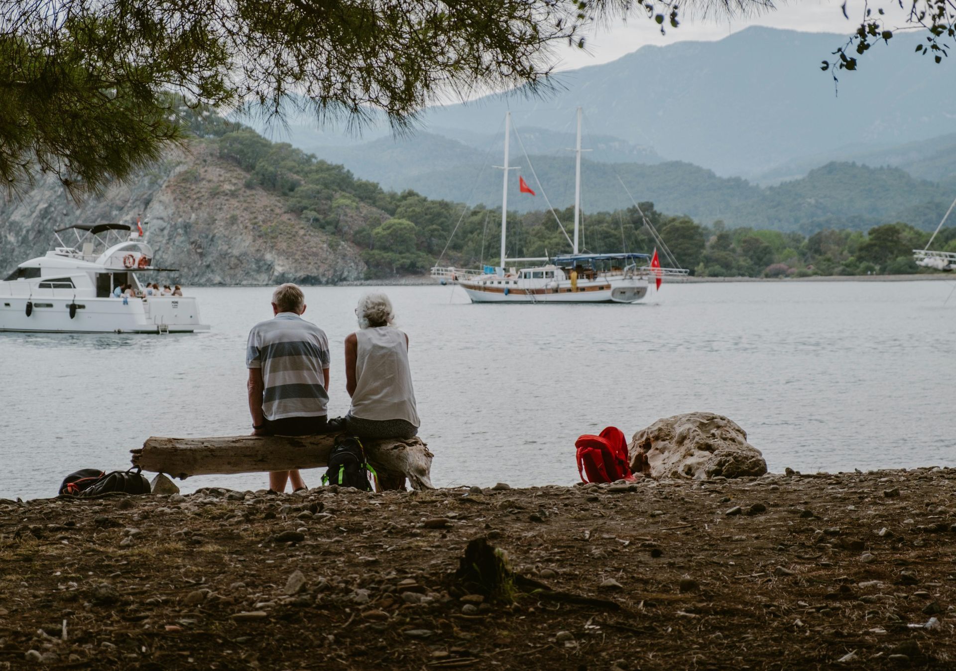 Two people sit on a wooden log by the water, looking at boats near a mountainous coastline.