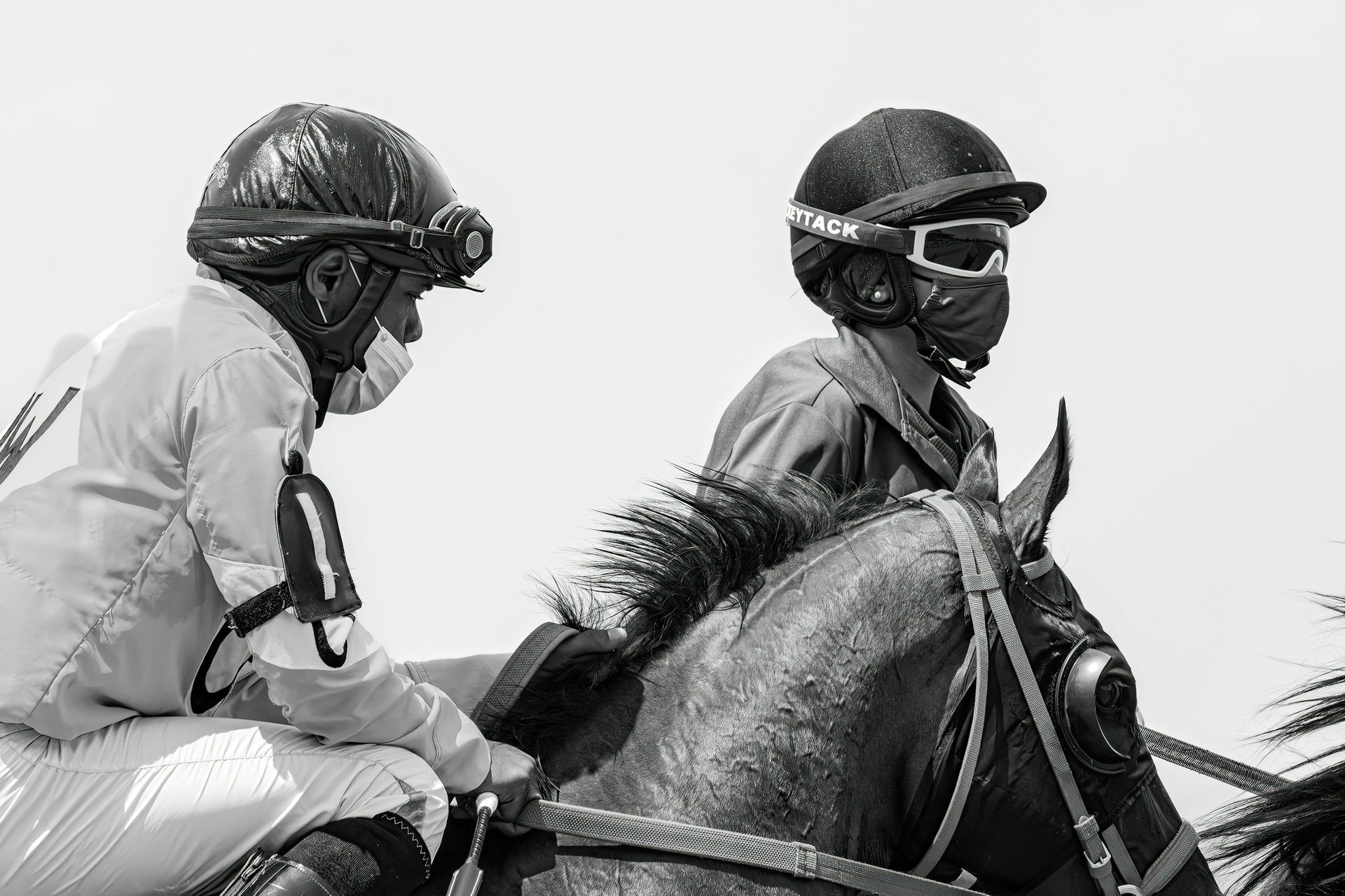 Two jockeys riding horses, wearing helmets and goggles, in a black-and-white race scene