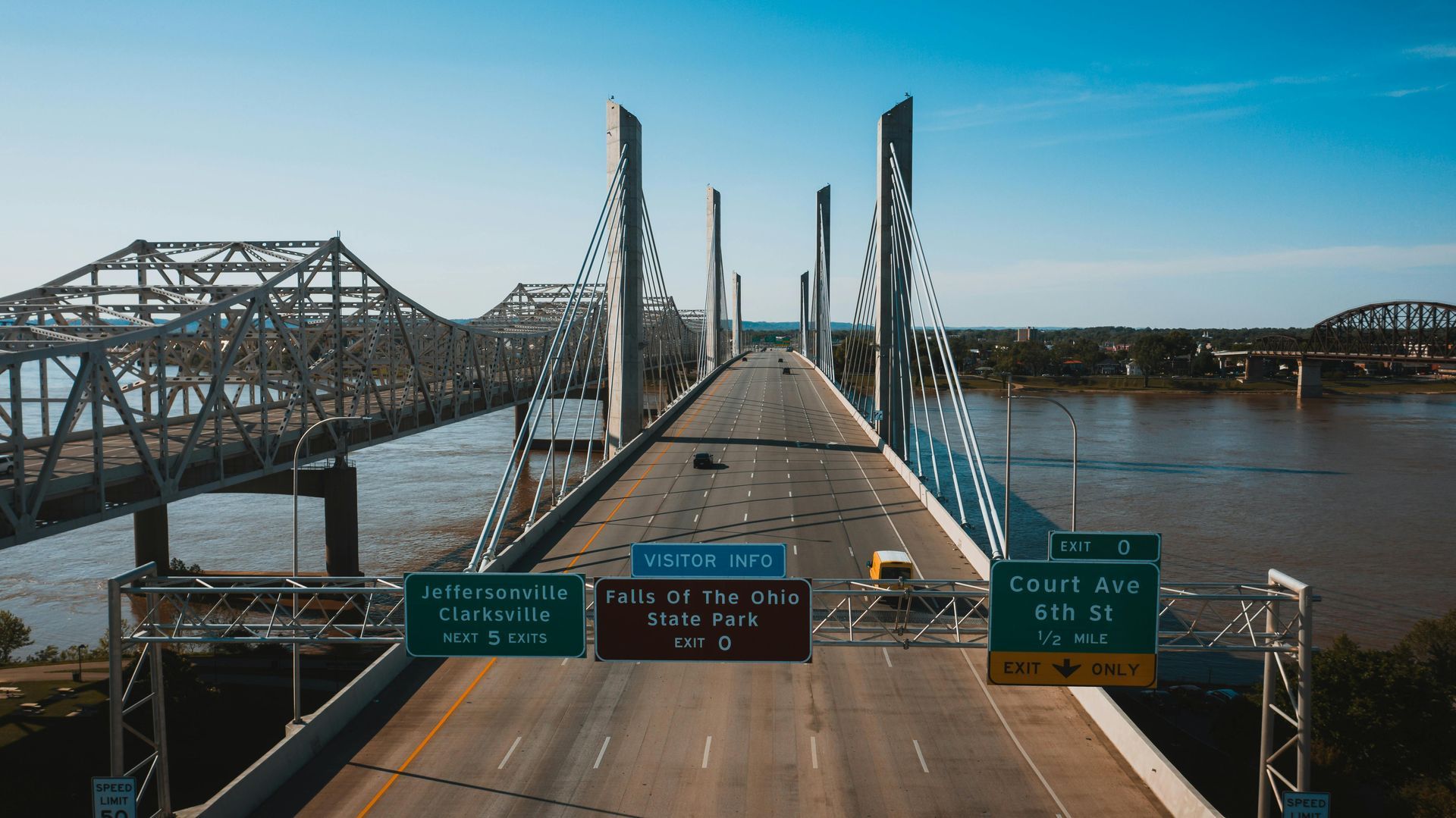 Bridges over a river, a cable-stayed bridge in focus, a truss bridge in the background, signage overhead, and a clear sky.
