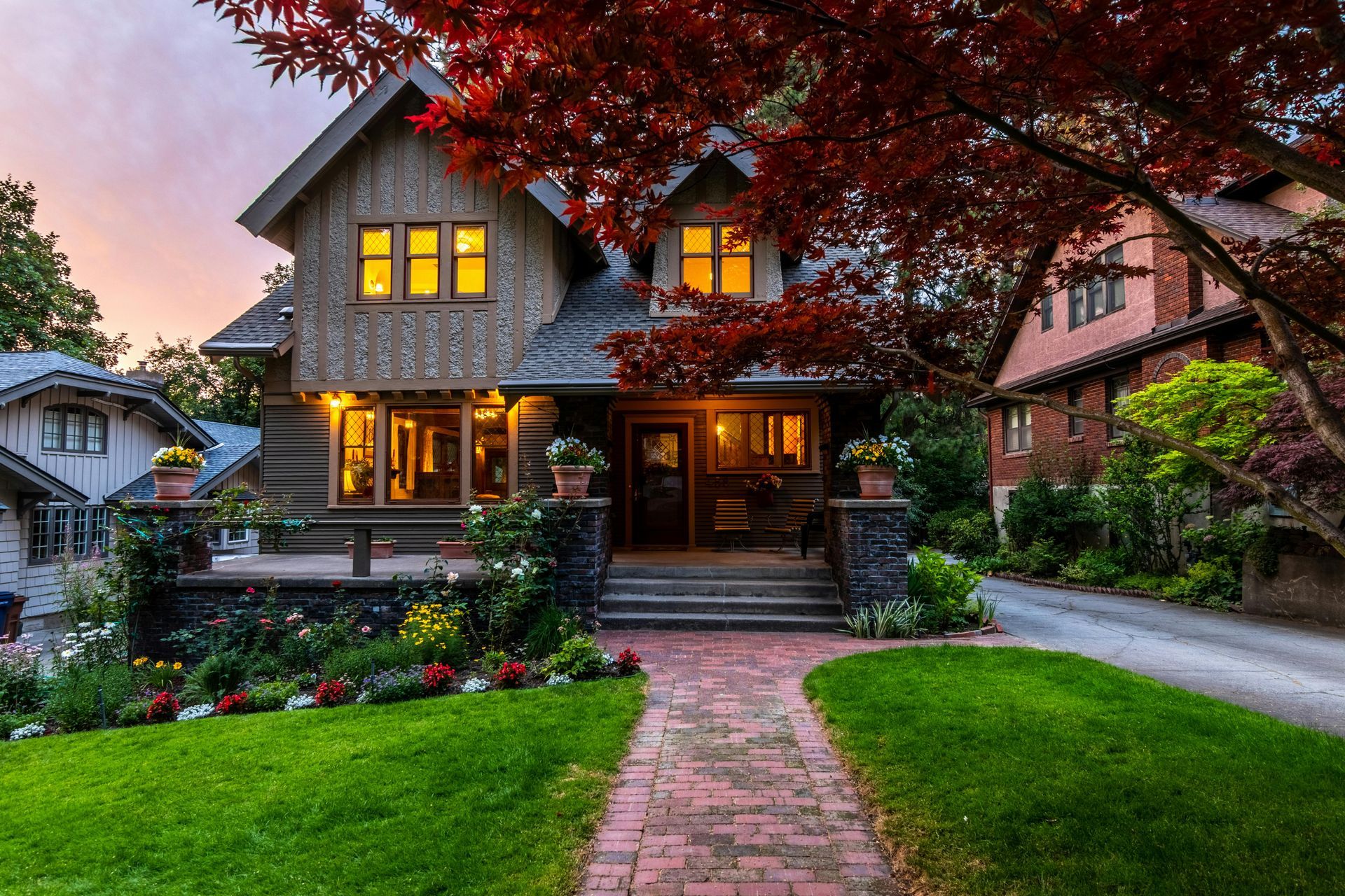 Charming Craftsman-style house with brick path, green lawn, red tree, and a driveway in the background.