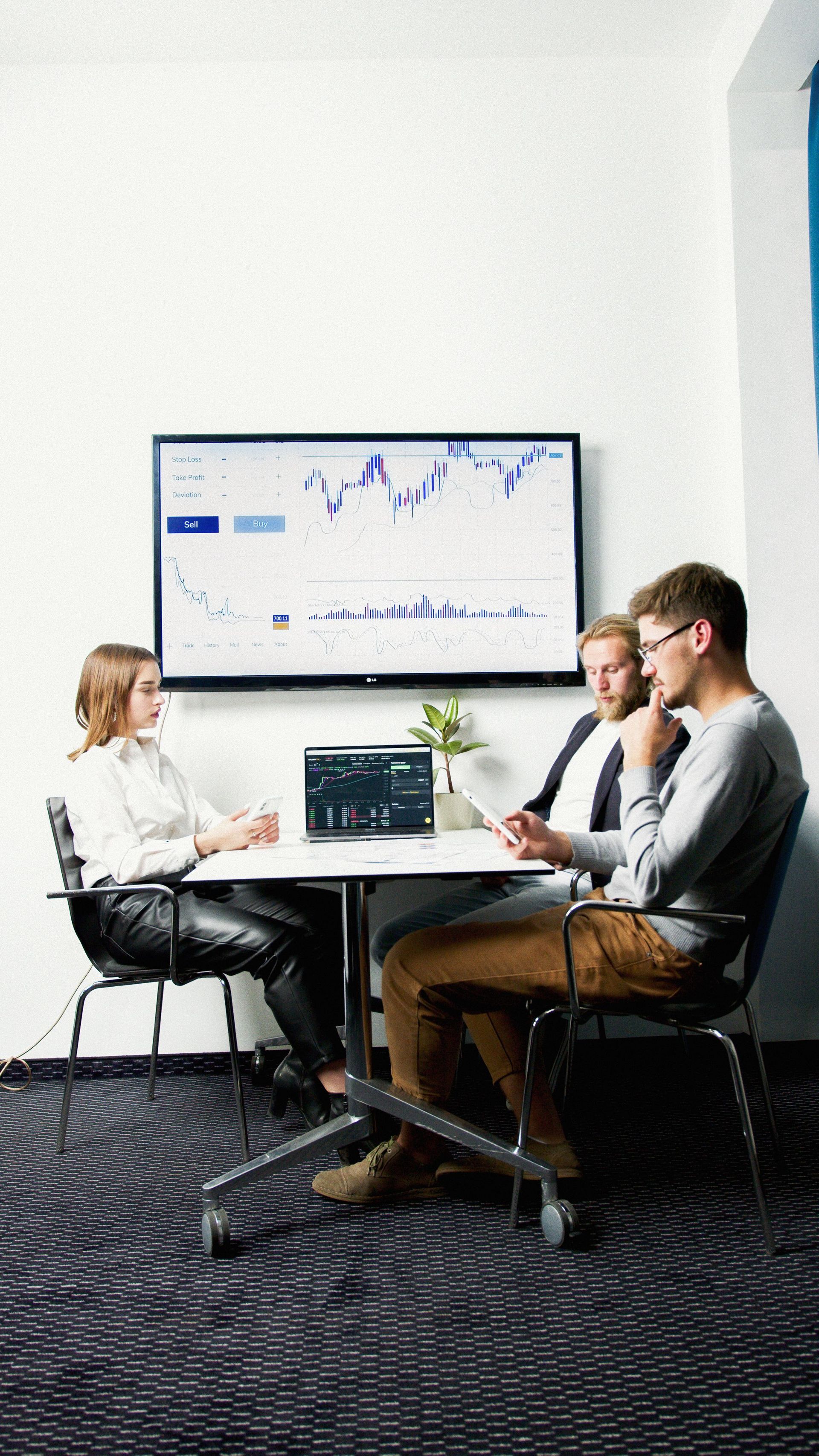 Three people in a meeting room discussing a presentation on a wall screen