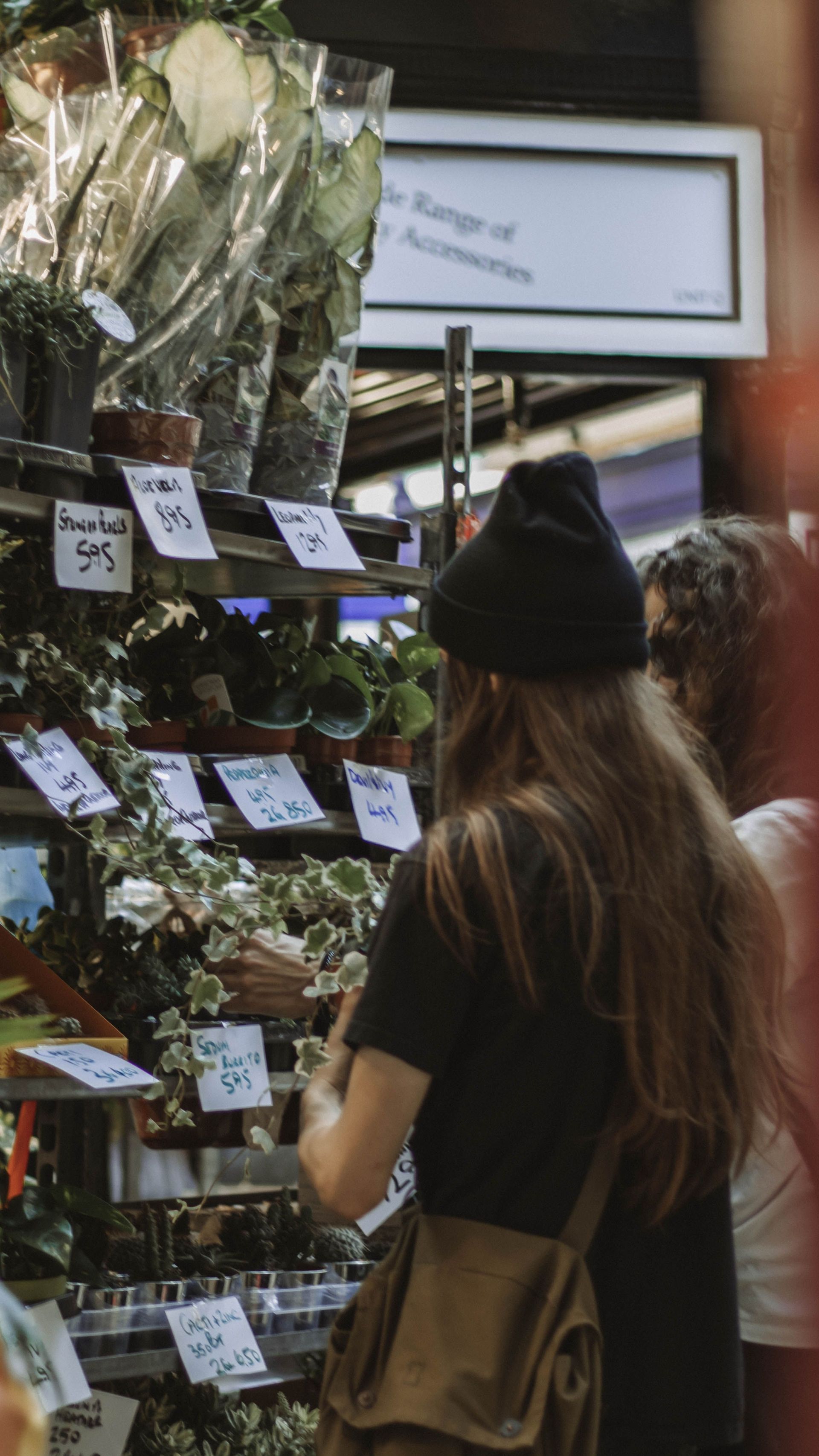 Person browsing flowers and plants at an outdoor market stall, seen from behind.