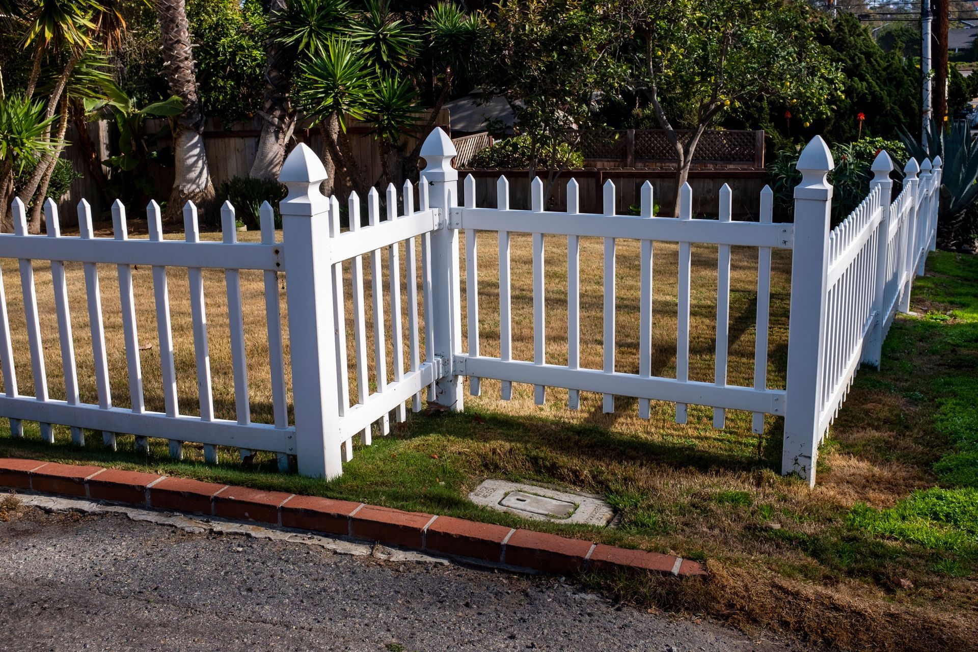 White picket fence enclosing a small grassy area; brick edging and trees in the background. White picket fence enclosing a small grassy area; brick edging and trees in the background.