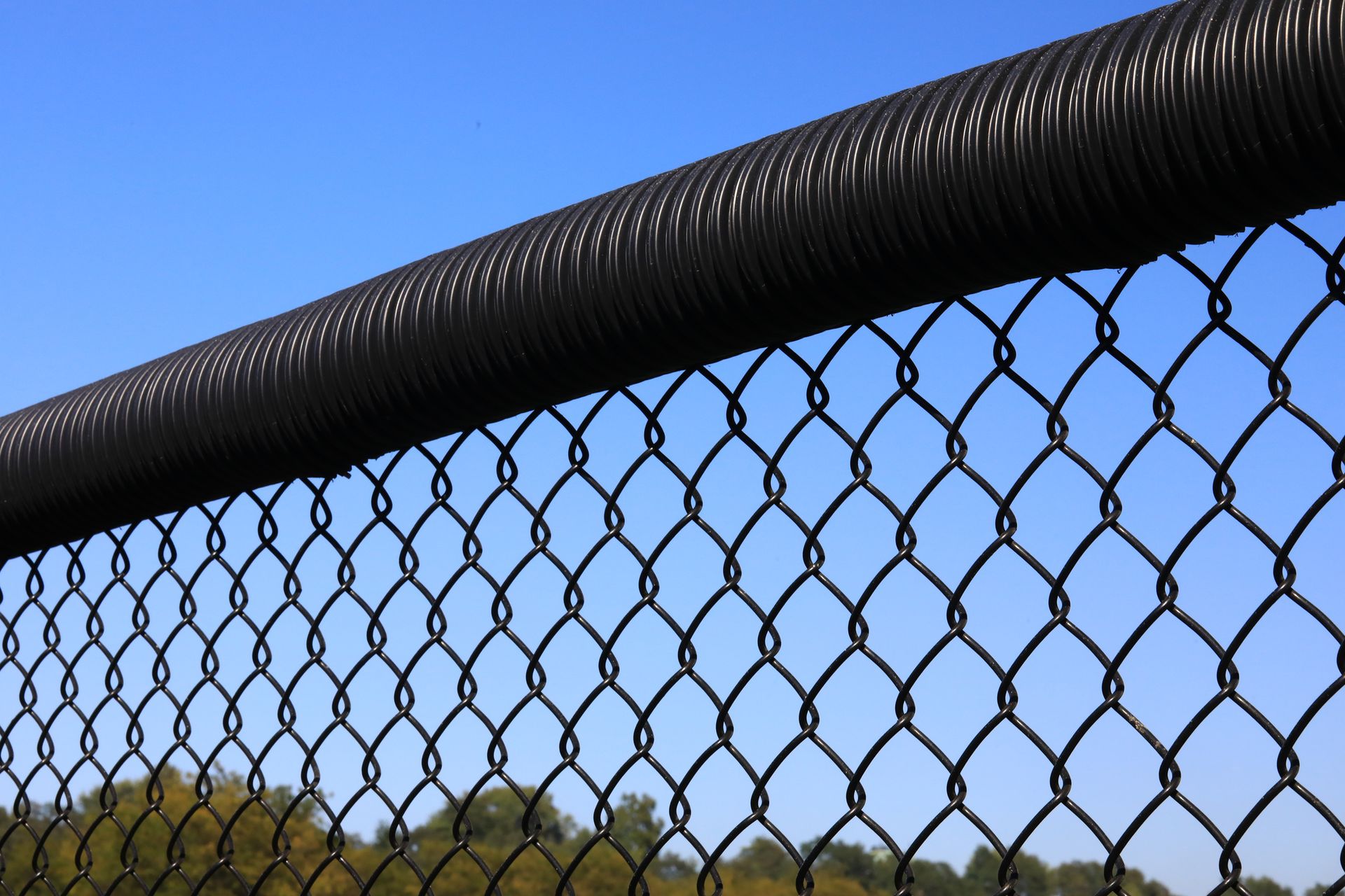 Black chain-link fence with black top rail against a bright blue sky, trees in the distance. Black chain-link fence with black top rail against a bright blue sky, trees in the distance.