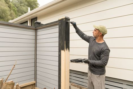 Man in gray sweater and cap painting a fence post black outside a house. Man in gray sweater and cap painting a fence post black outside a house.
