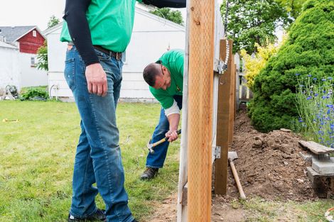 Two people building a wooden fence outdoors; one hammers, the other steadies the post. Two people building a wooden fence outdoors; one hammers, the other steadies the post.