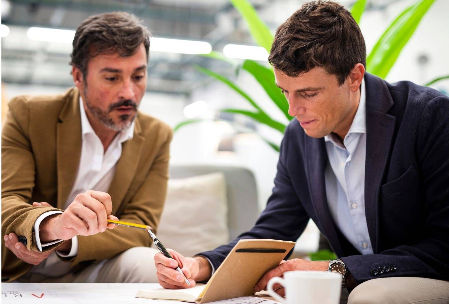 Two men in blazers review notes at a table with a tablet and coffee cup