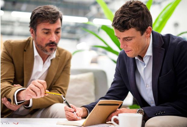 Two men in blazers review documents and write together at a table in a bright office
