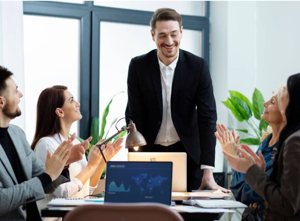 Team applauding a smiling presenter in a bright conference room with laptops and plants